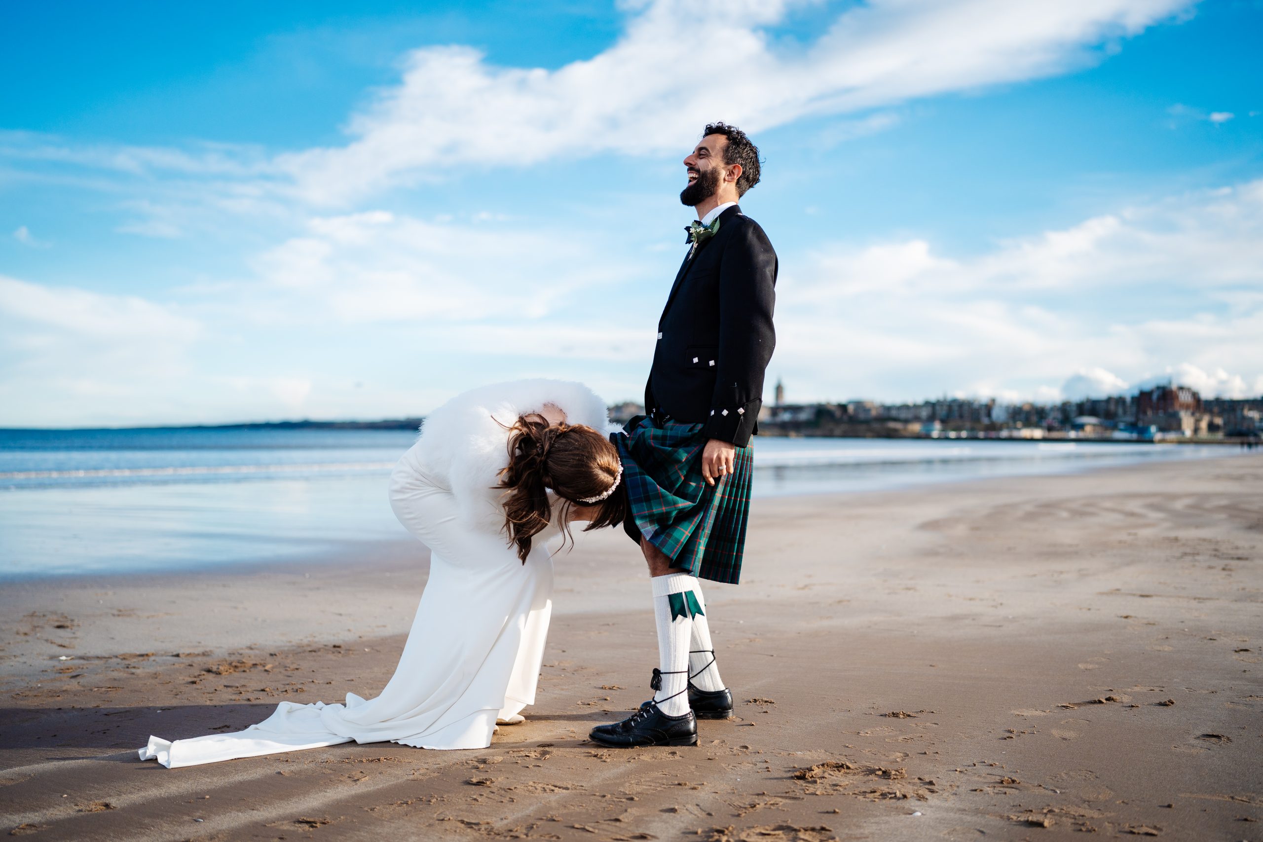 Bride cheekily peeks under groom's kilt whilst on St Anrew's West Sands beach on their wedding day