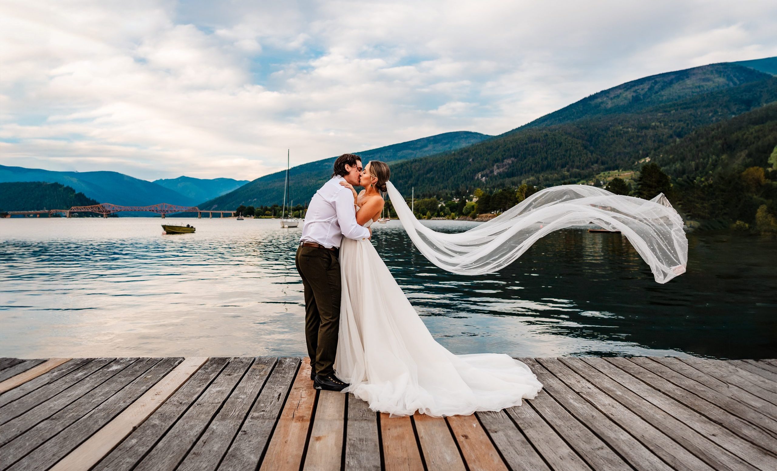Bride and groom embrace on a pier over a lake in Nelson, British Columbia, the Brides veil blows in the wind