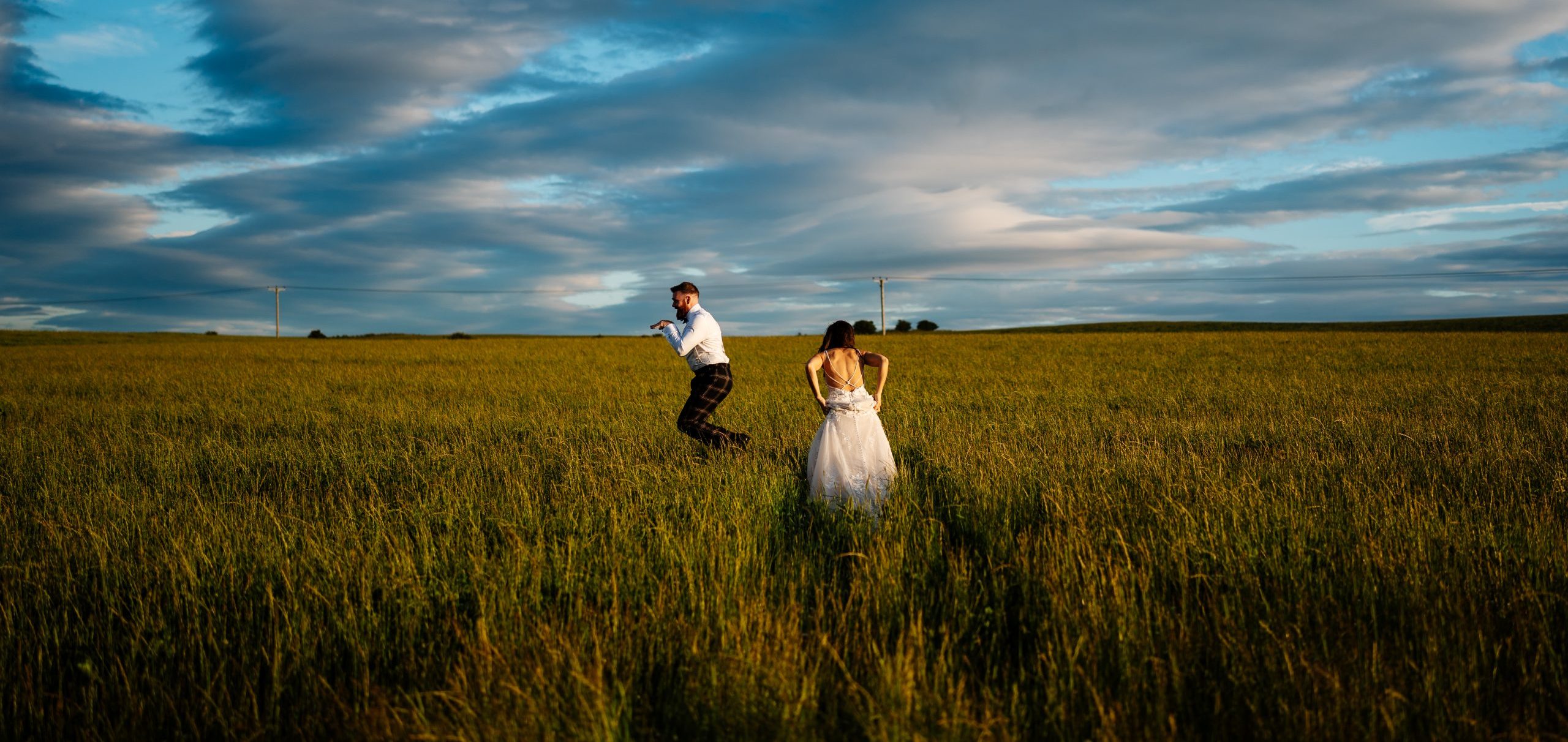 Groom leaping through a field like a bunny, whilst his wife walks through the fields at Falside Mill